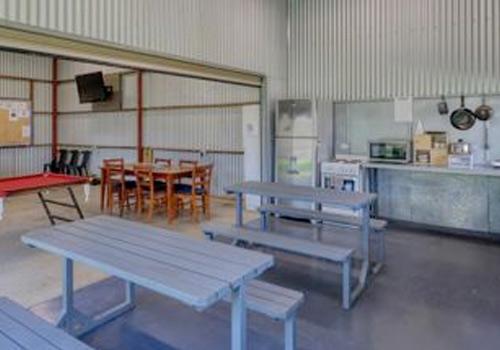 A spacious indoor community area at Barham Caravan Park, featuring picnic tables, benches, a ping pong table, and a kitchenette with appliances and utensils.