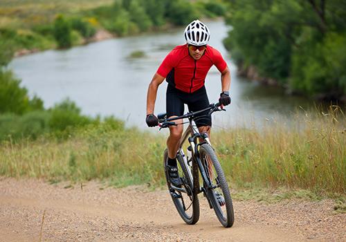 A cyclist in a red jersey and helmet rides along a gravel path near a serene river