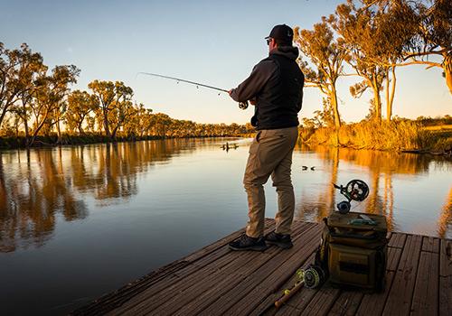A man fishing on a wooden dock at sunset, casting his line into calm waters surrounded by trees and golden light.
