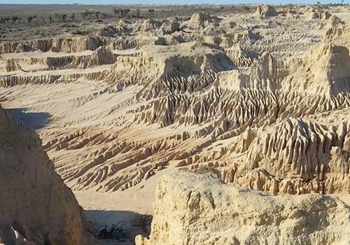 A rugged, sunlit landscape featuring layered rock formations and deep erosion patterns, showcasing the raw beauty of a desert canyon.