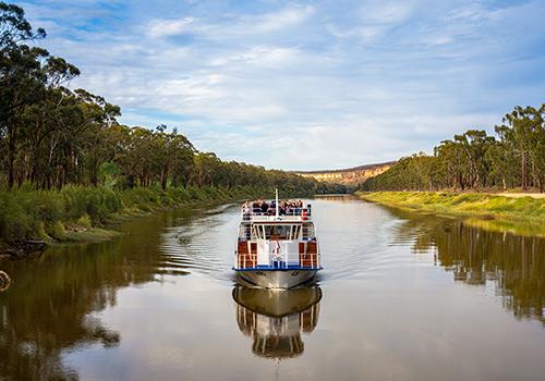 A scenic river cruise boat glides through calm waters, surrounded by lush greenery and a picturesque landscape under a partly cloudy sky.