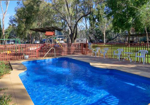 A bright blue swimming pool surrounded by a red fence, with lounge chairs and a shaded seating area under a wooden pergola, set against a backdrop of lush green trees.
