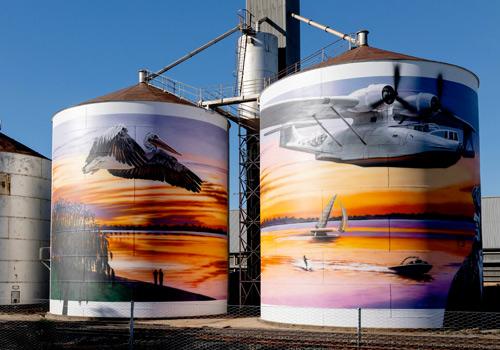Two large industrial tanks painted with vibrant murals depicting a pelican in flight over a sunset and a seaplane soaring above a sailboat on the water.