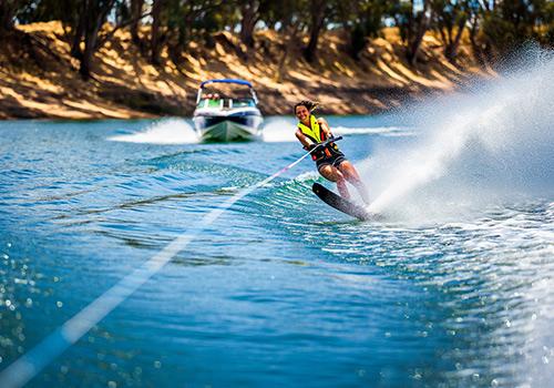 A thrilling water skiing scene on the Murray River, with a person gliding across the water behind a boat, surrounded by calm blue waters and lush green trees along the riverbank.